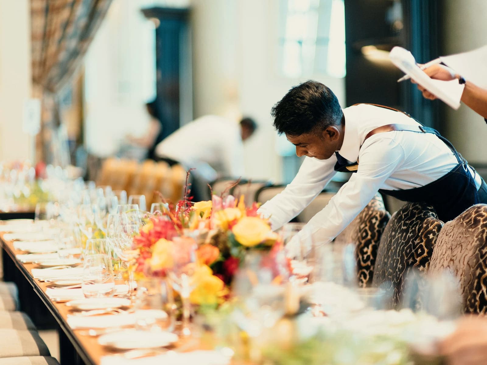 A member of event staff setting a long banquet table with floral centerpieces