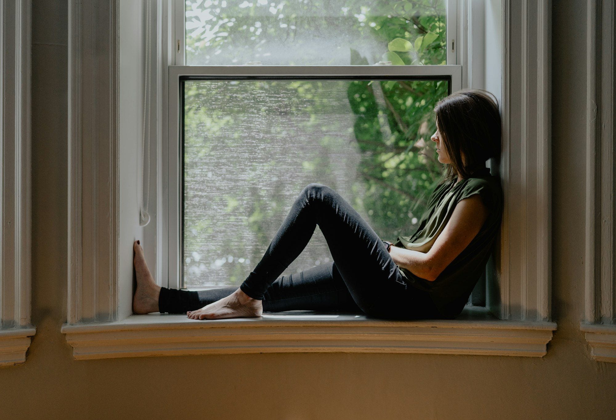 A person pausing by a window with their eyes closed, taking a slow breath